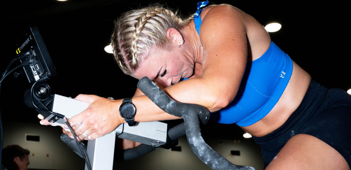 A woman in a blue sports bra and black shorts riding a stationary exercise bike in a dark gym setting.