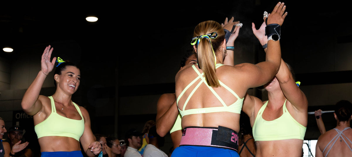 Three women in matching athletic wear—light yellow sports bras and blue shorts—high-fiving in a gym or event space. One woman faces away from the camera, showing a pink and black waist belt, while the others face toward the camera.